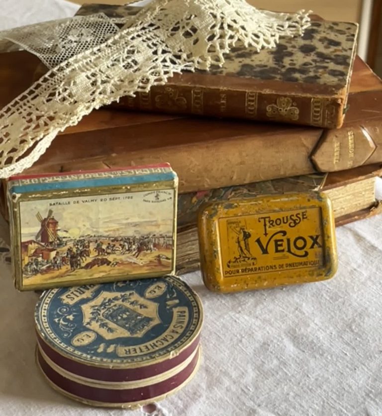 Livres et dentelles Livres anciens, boîtes métalliques vintage, et dentelle ancienne, posés sur un tissu clair